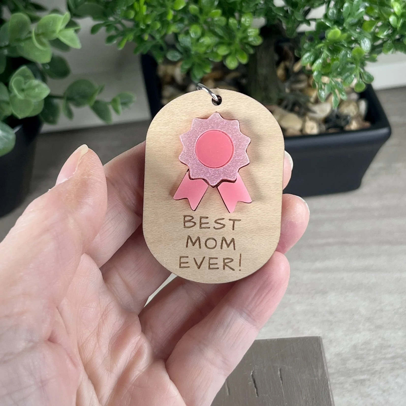 handheld laser cut maple wood keychain featuring glimmering/gloss pink acrylic ribbon and engraved with the phrase "BEST MOM EVER!".