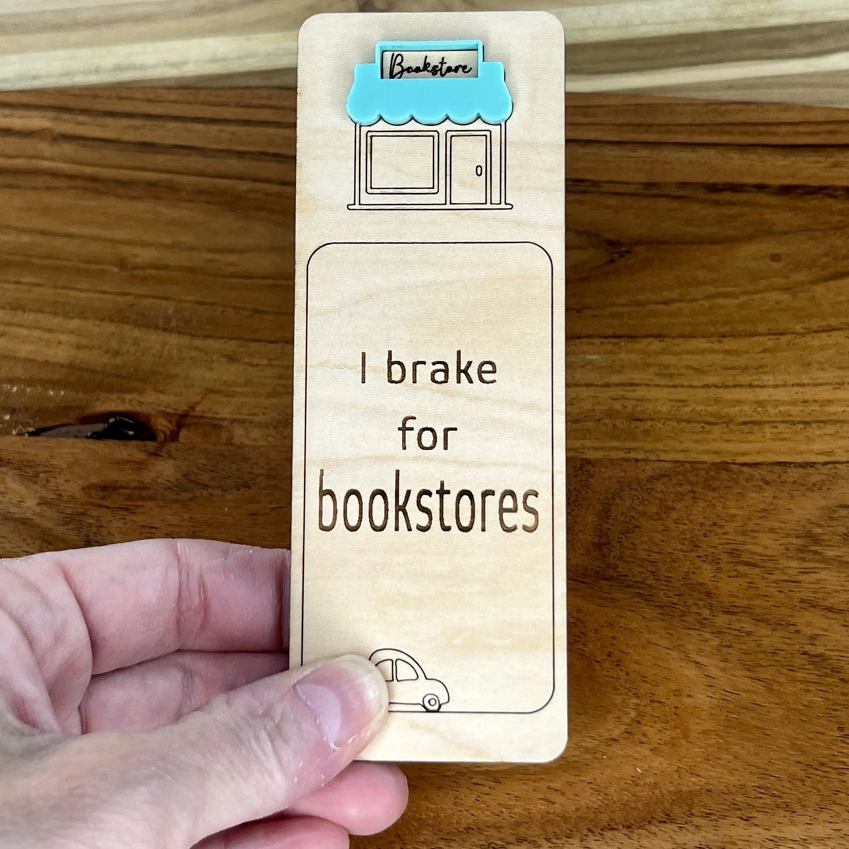 handheld maple bookmark featuring mint acrylic bookstore awning above a bookstore storefront illustration engraved with the phrase "I brake for bookstores".