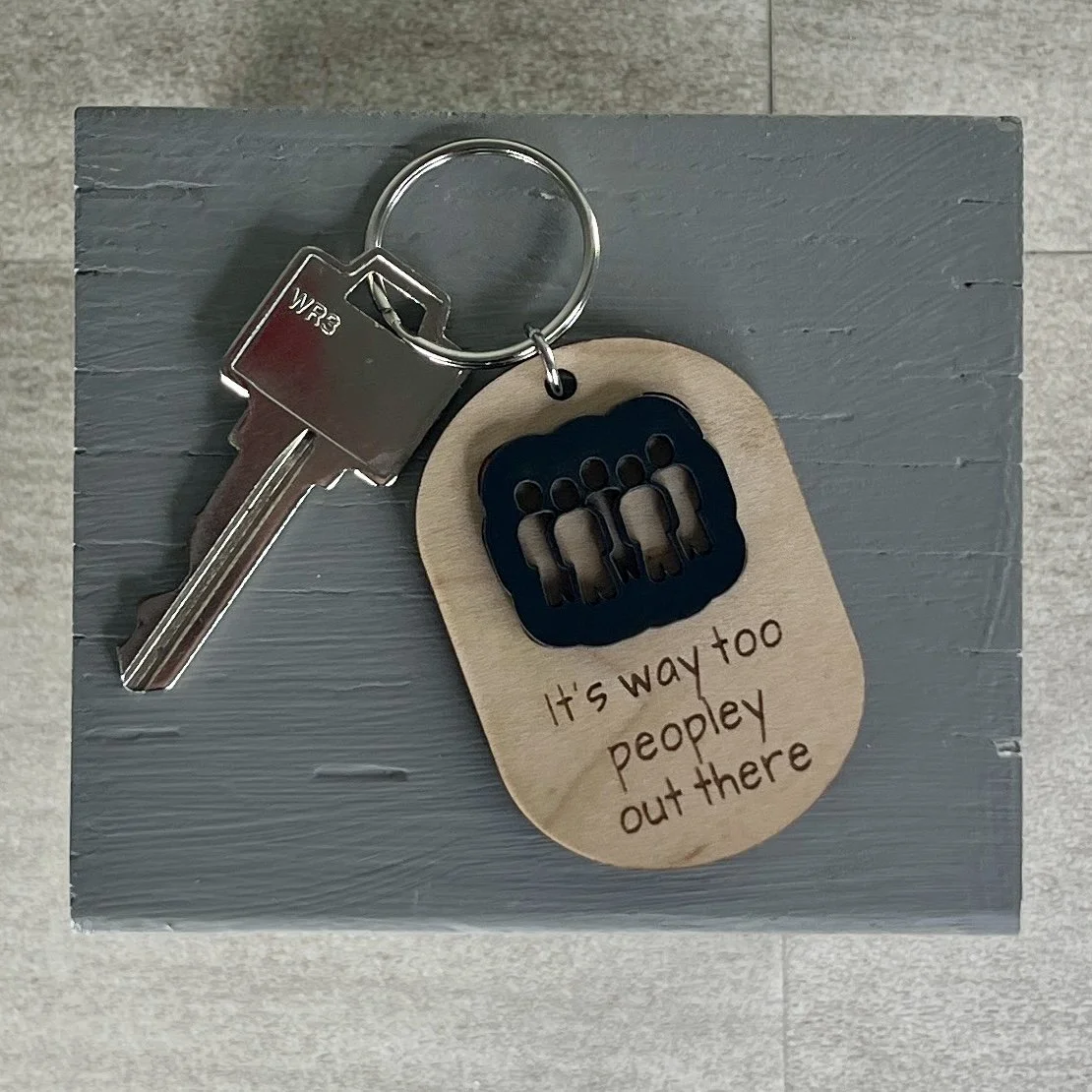 lifestyle shot of laser cut maple wood keychain with silhouette of group of people in matte black acrylic and the engraved phrase "It's way to peopley out there".