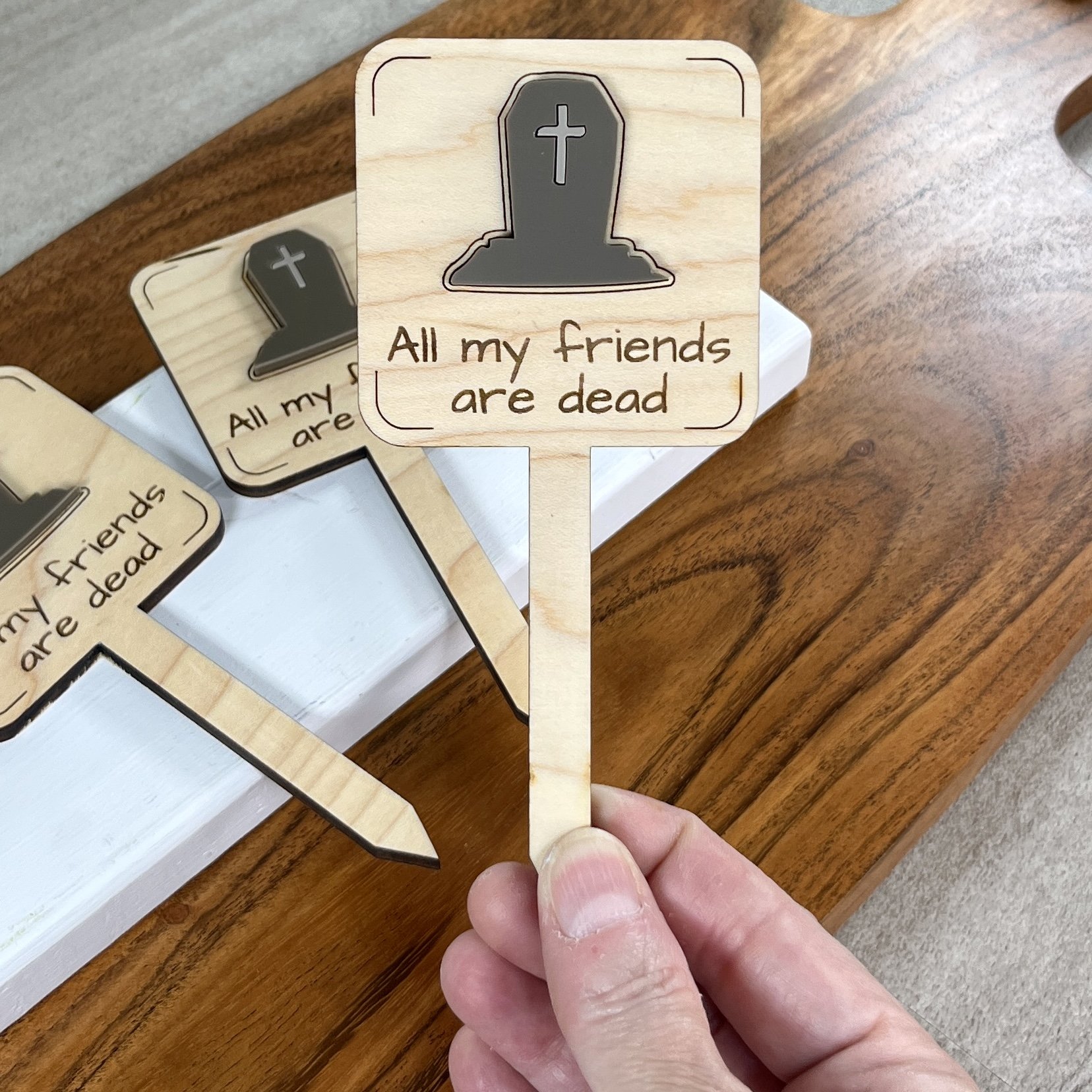 handheld maple plant stake featuring an acrylic espresso tombstone embellishment engraved with the phrase "All my friends are dead".
