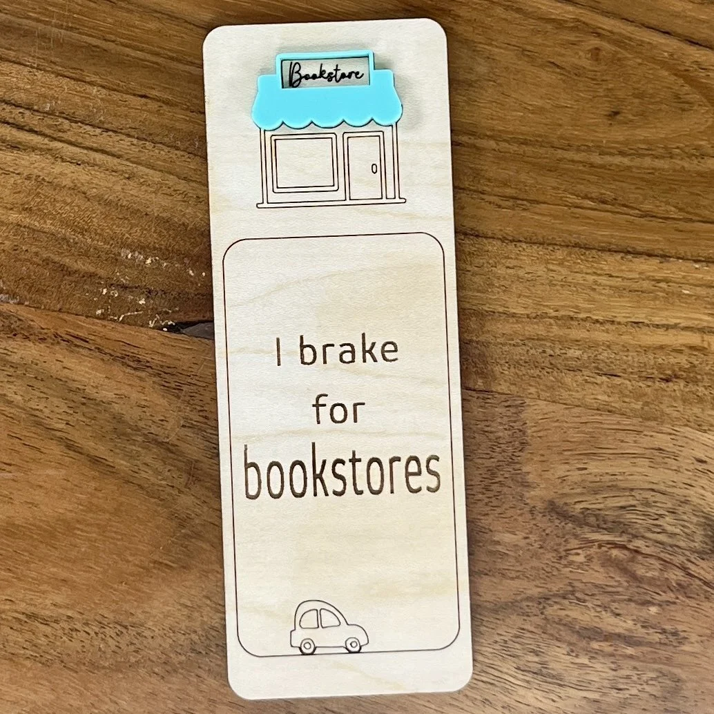 flat lay maple bookmark featuring mint acrylic bookstore awning above a bookstore storefront illustration engraved with the phrase "I brake for bookstores".