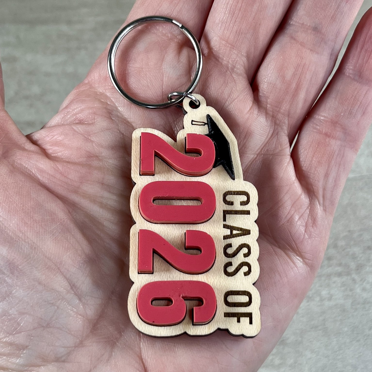 Hand holding a Class Of graduation keychain made from engraved maple wood with raised matte acrylic numbers and a small black graduation cap at the top.
