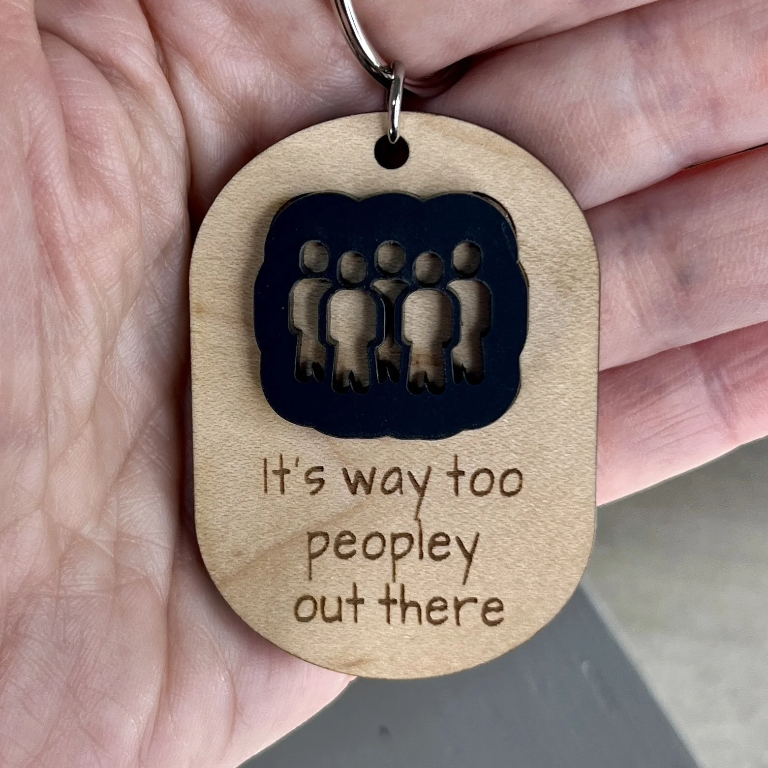 handheld laser cut maple wood keychain with silhouette of group of people in matte black acrylic and the engraved phrase "It's way to peopley out there".