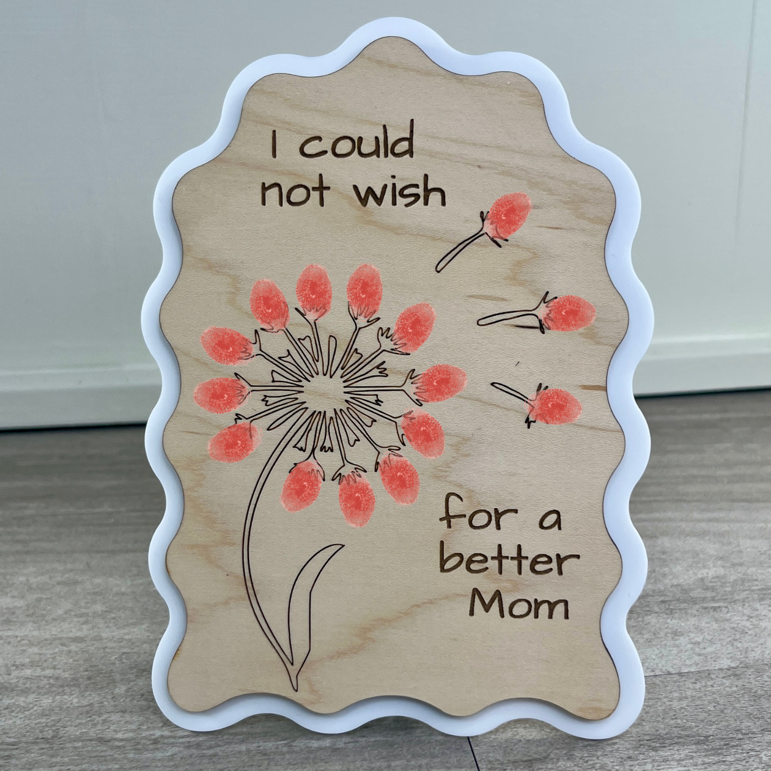 Maple wood sign in a wavy-edged rectangle with a white acrylic border, engraved with a dandelion and the message "I could not wish for a better Mom," with a child's painted fingerprints as petals.