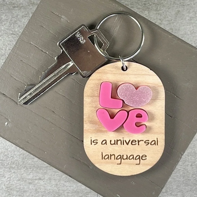 lifestyle shot of laser cut maple wood keychain featuring layered pink acrylic letters spelling "Love" with heart details and the engraved phrase "is a universal language."