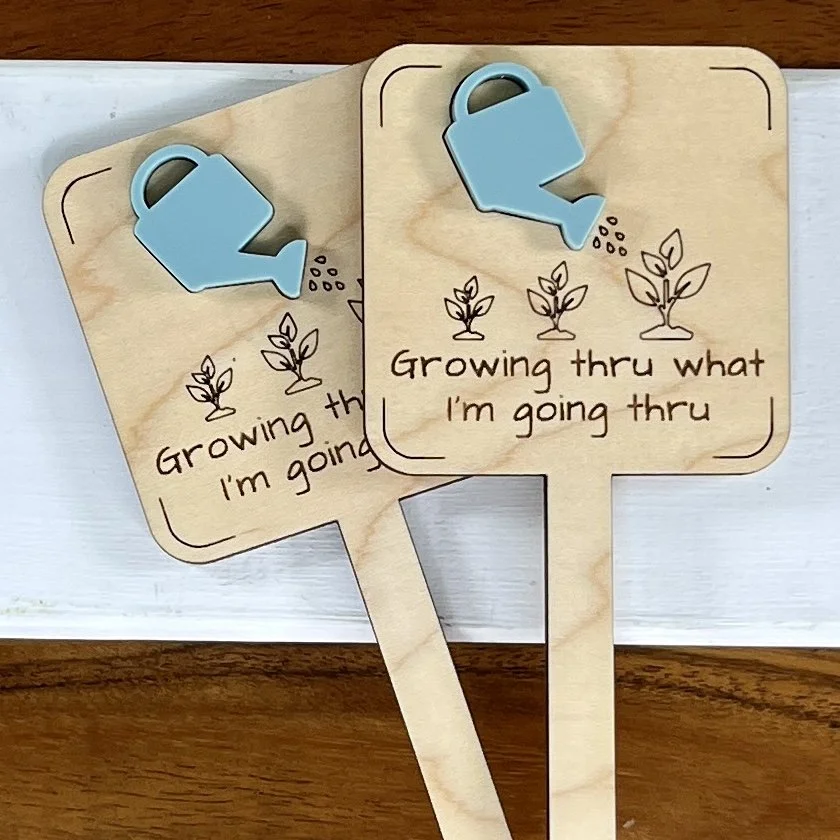 flat lay maple plant stake featuring an acrylic watering can embellishment and engraved plant growth design with the phrase "Growing Thru What I'm Going Thru".