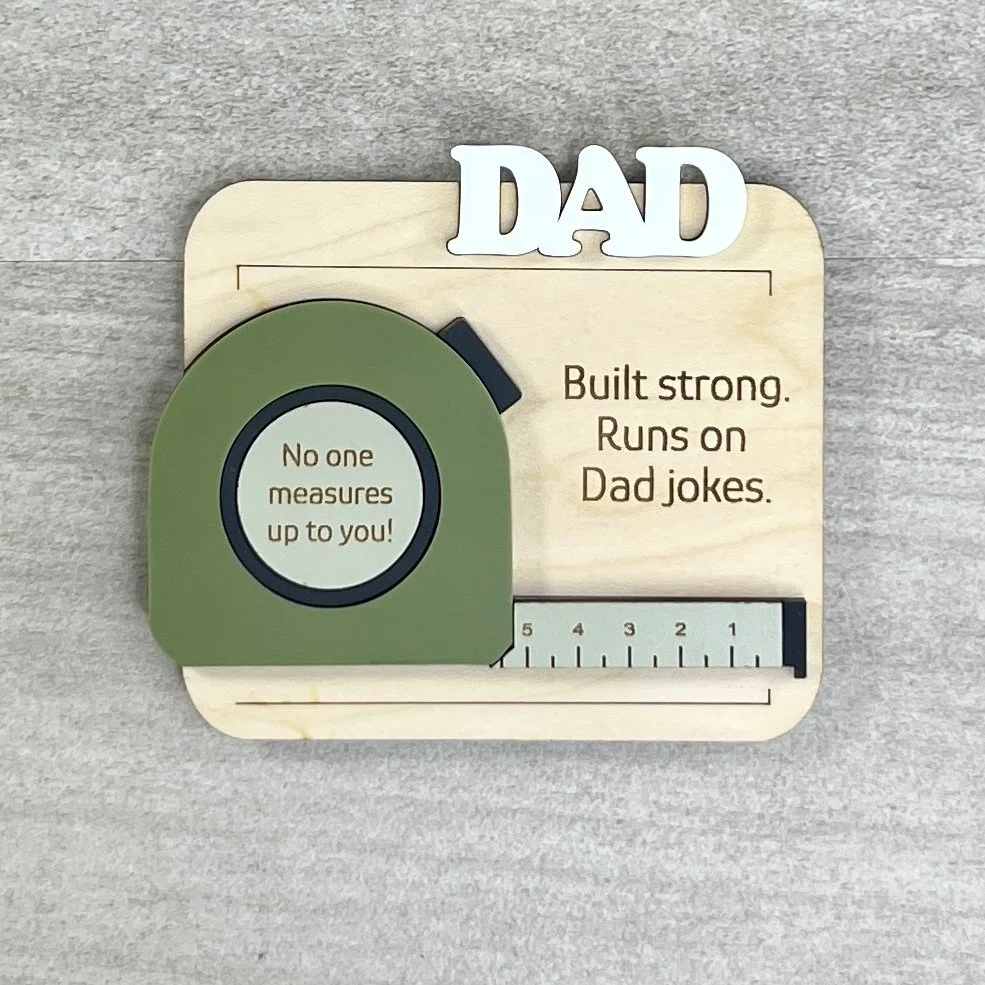 Laser cut maple magnet with raised olive acrylic tape measure design. Header features raised white text. Engraved with quotes "Built strong.  Runs on Dad jokes." and "No one measures up to you".