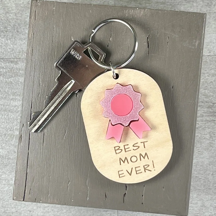 lifestyle shot of laser cut maple wood keychain featuring glimmering/gloss pink acrylic ribbon and engraved with the phrase "BEST MOM EVER!".