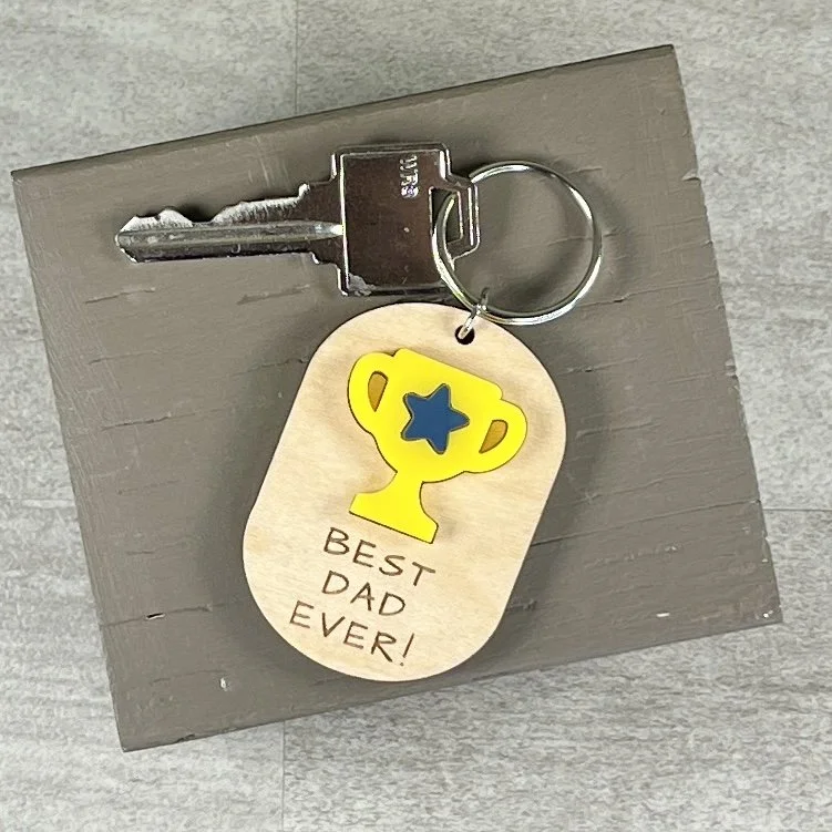 lifestyle shot of laser cut maple wood keychain featuring matte bumblebee yellow acrylic trophy decorated with a navy blue acrylic star and the engraved phrase "BEST DAD EVER!"