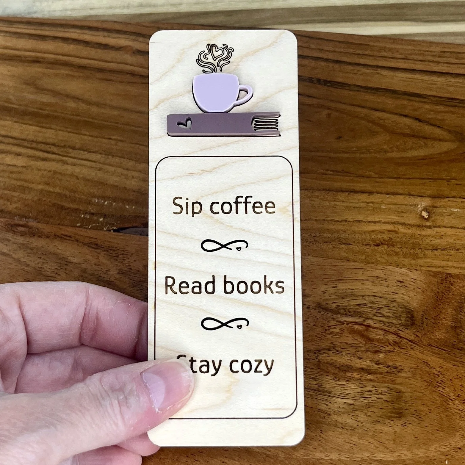 handheld maple bookmark featuring an ash rose coffee cup and mauve book embellishment engraved with the phrase "Sip coffee Read books Stay cozy".