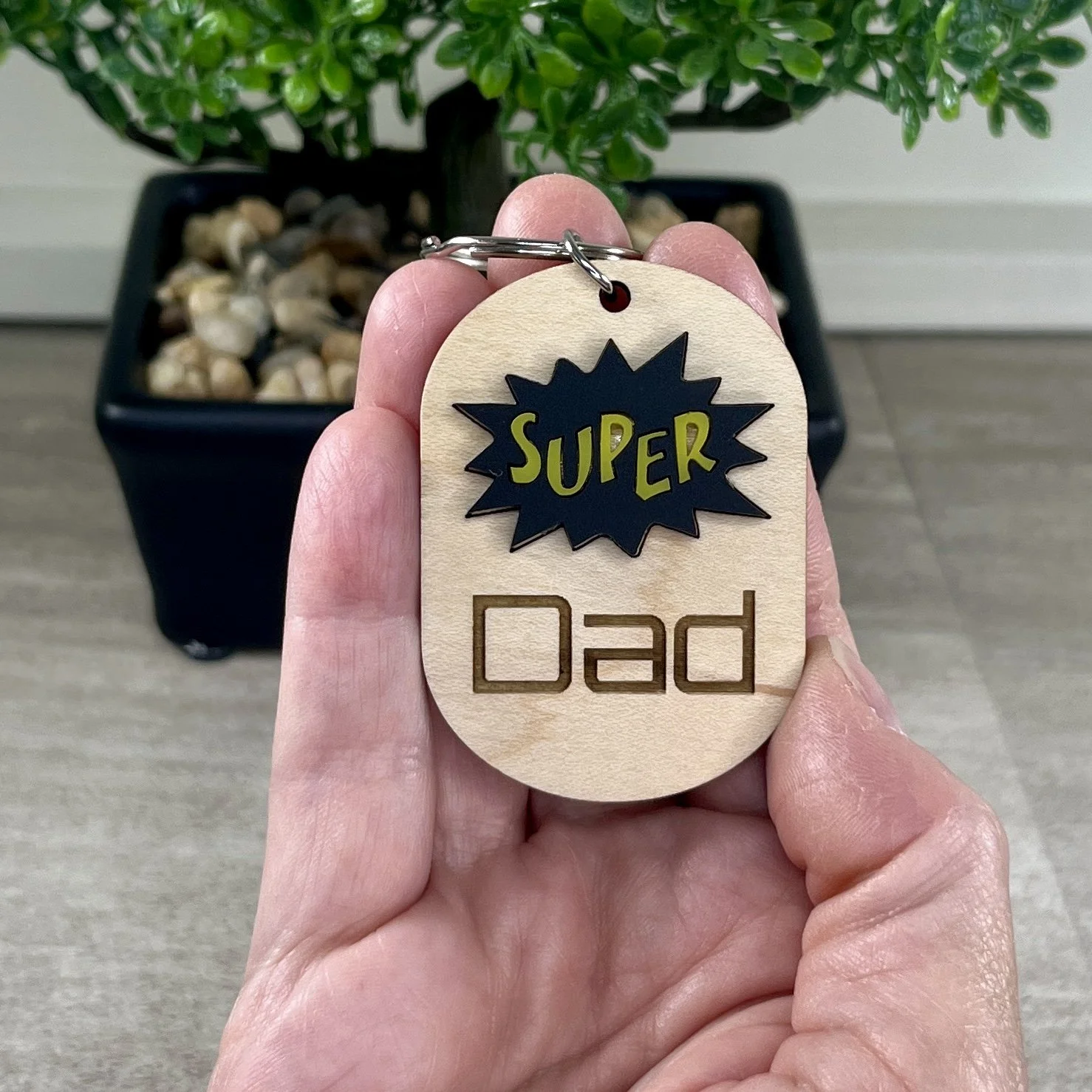 handheld view wood keychain black and yellow comic book burst shape reading Super with Dad engraved beneath.