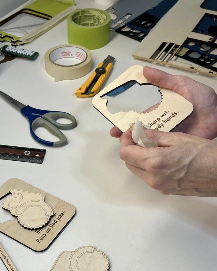 Person holding a wooden beer mug cutout with the phrase "Sharp wit...” on a worktable with crafting tools, tape, scissors, ruler, and other wooden cutouts.