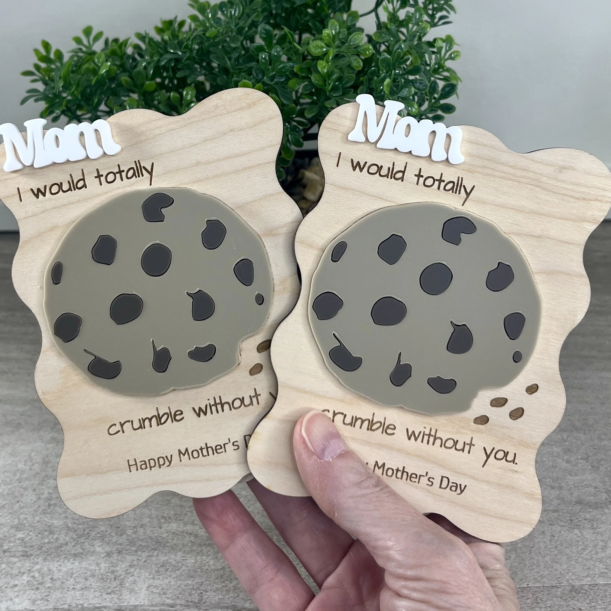 two handheld laser cut wavy-edged maple magnets featuring the word Mom in raised white acrylic, a chocolate chip cookie in acrylic and engraved with the phrase "I would totally crumble without you".