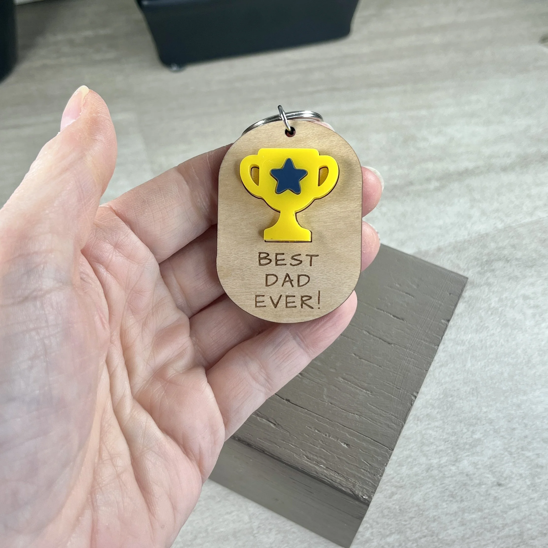 handheld laser cut maple wood keychain featuring matte bumblebee yellow acrylic trophy decorated with a navy blue acrylic star and the engraved phrase "BEST DAD EVER!"