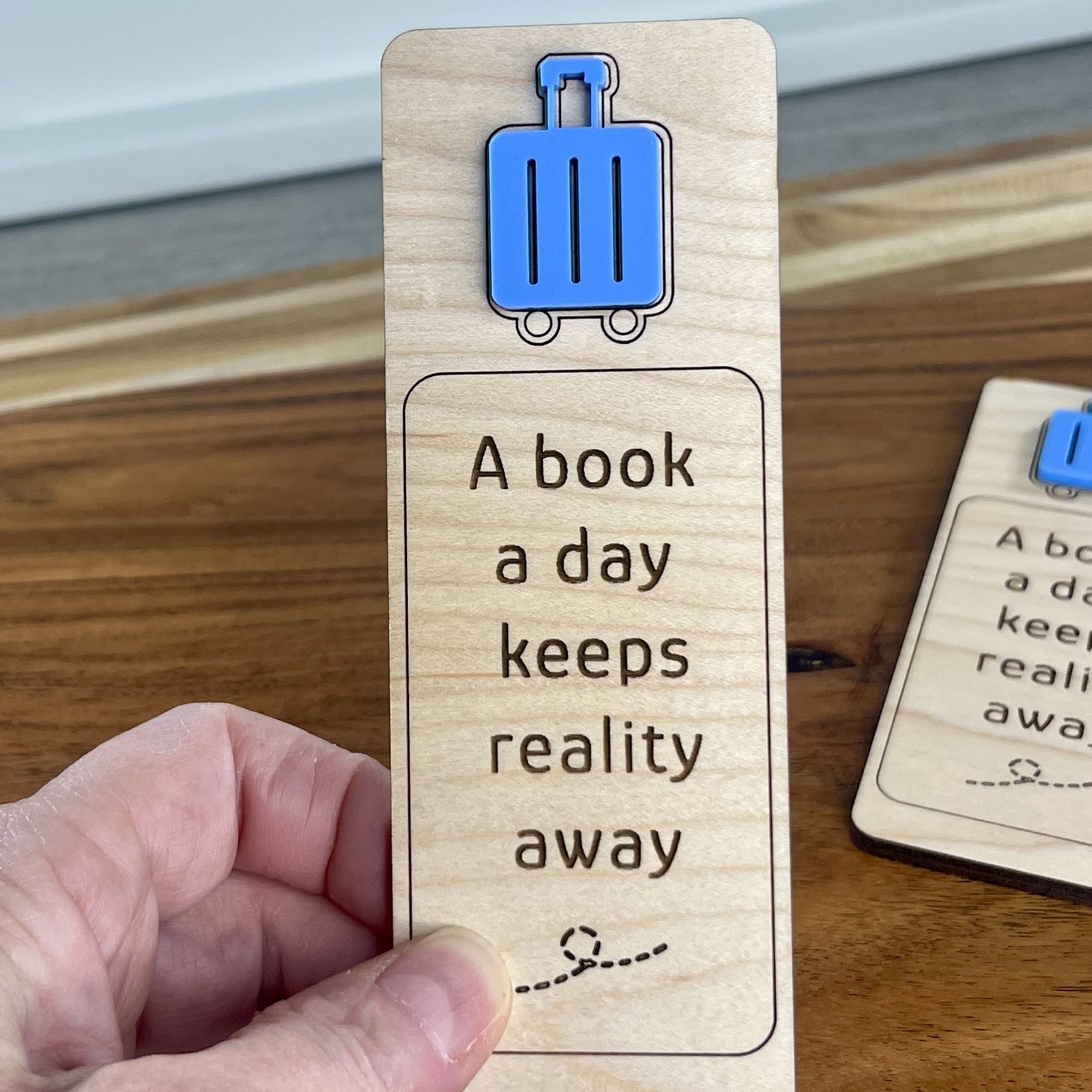 handheld maple bookmark showing blue acrylic suitcase embellishment and engraved phrase "A book a day keeps reality away".