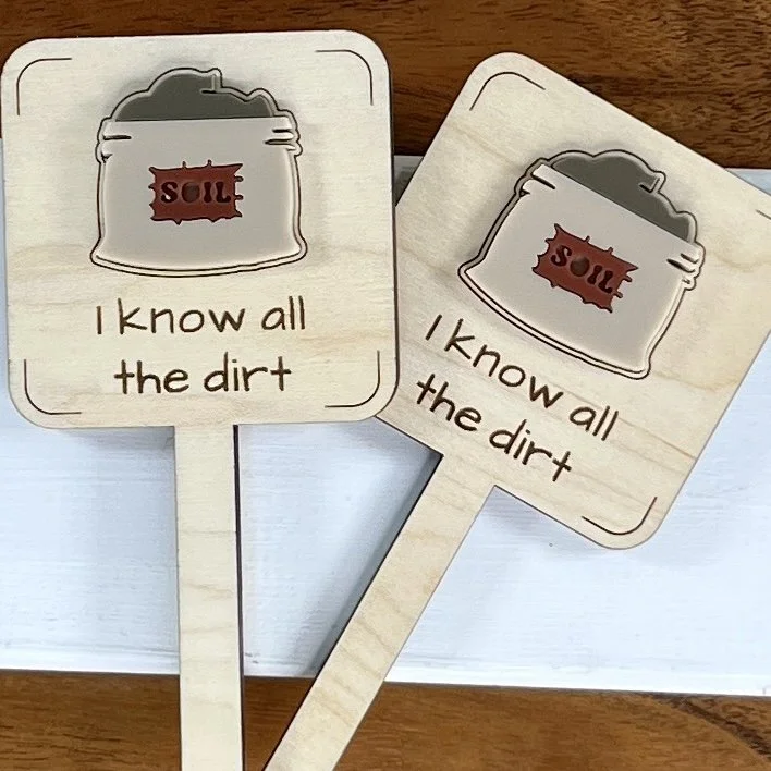 flat lay maple plant stake featuring an acrylic bag of soil embellishment engraved with the phrase "I know all the dirt".