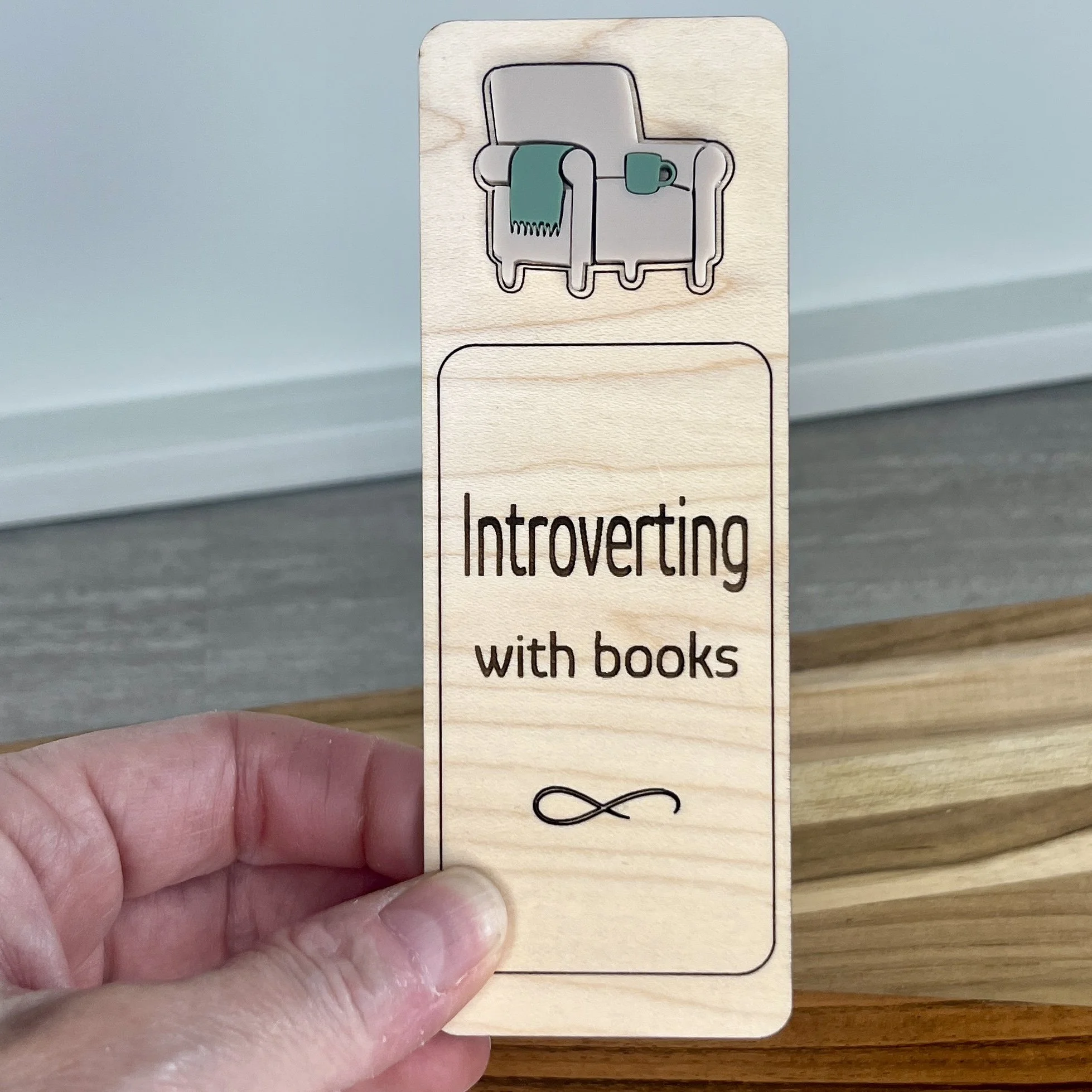 handheld maple bookmark featuring a comfy sand coloured acrylic chair with sage blanket and coffee mug embellishment engraved with the phrase "Introverting with books".