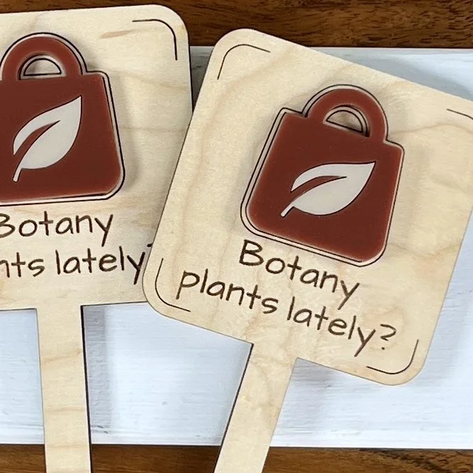 flat lay maple plant stake featuring an acrylic garden centre shopping bag embellishment engraved with the phrase "All my friends are dead".