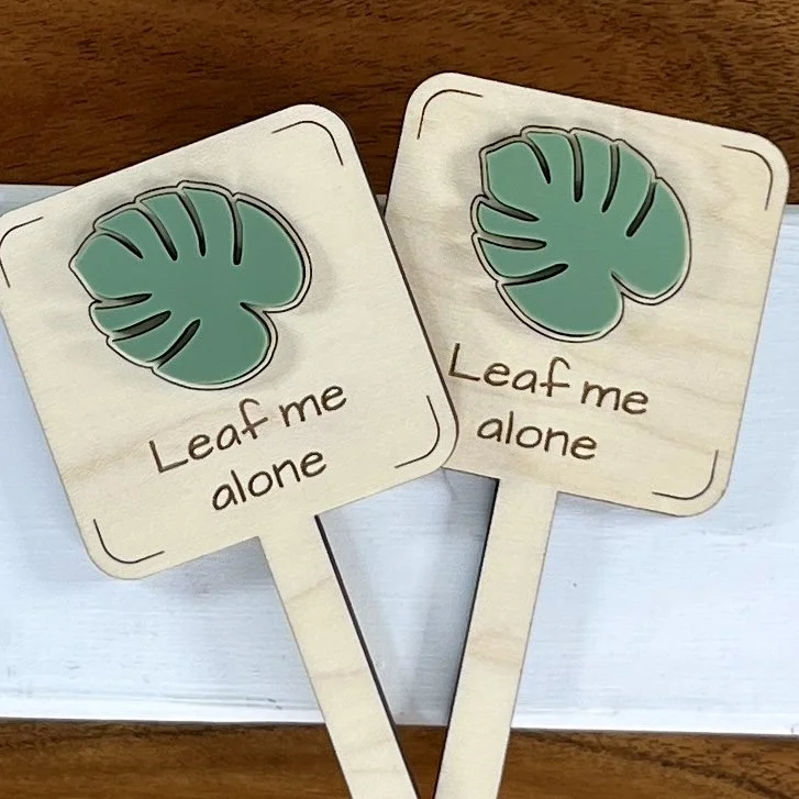 flat lay maple plant stake featuring a monstera leaf embellishment engraved with the phrase "Leaf me alone".