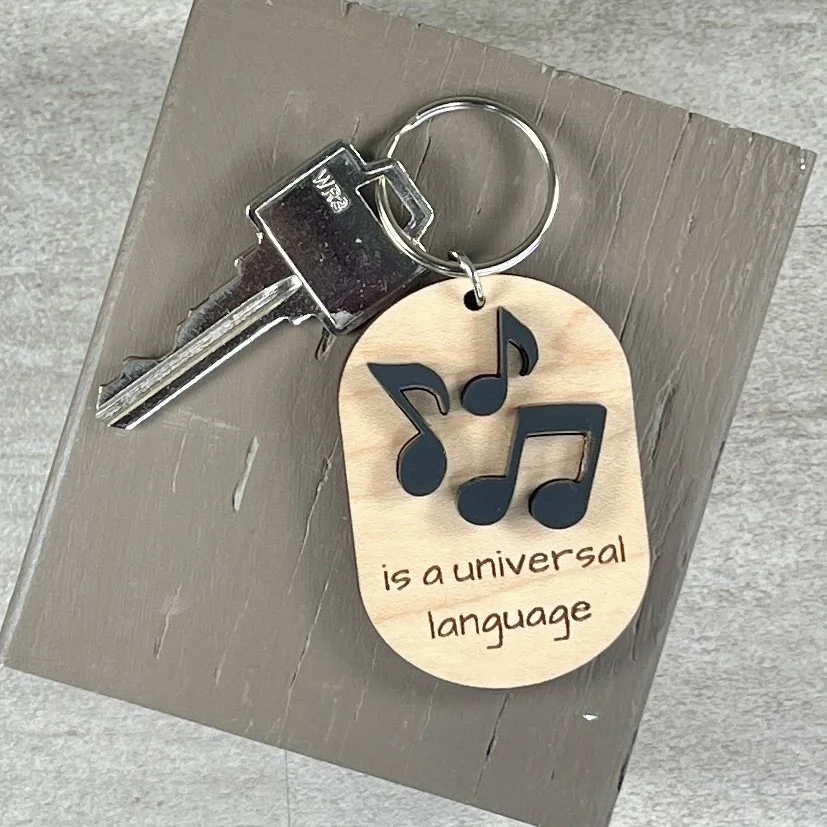 lifestyle shot of laser cut maple wood keychain featuring three raised musical notes in matte black acrylic and the engraved phrase "is a universal language."