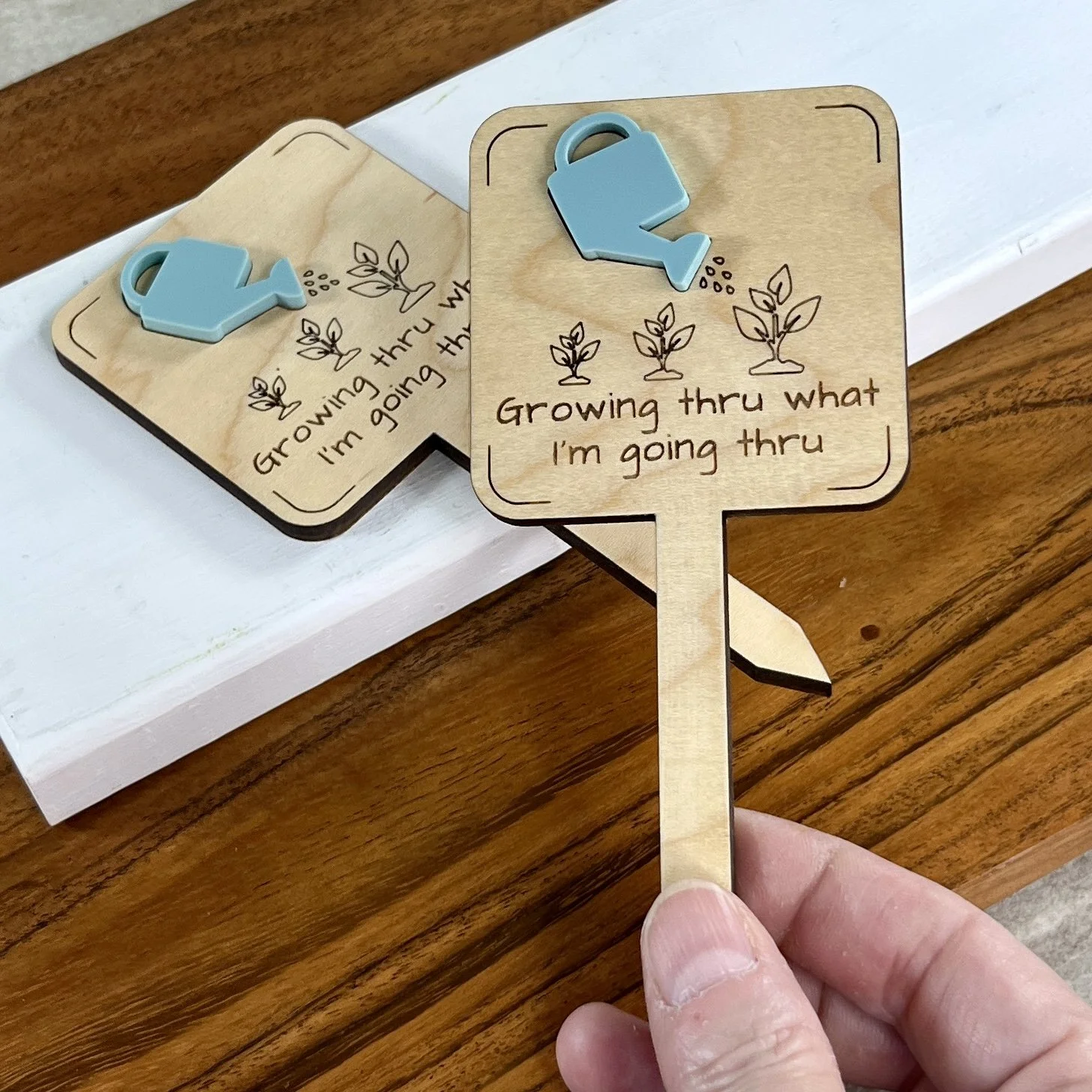 handheld maple plant stake featuring an acrylic watering can embellishment and engraved plant growth design with the phrase "Growing Thru What I'm Going Thru".