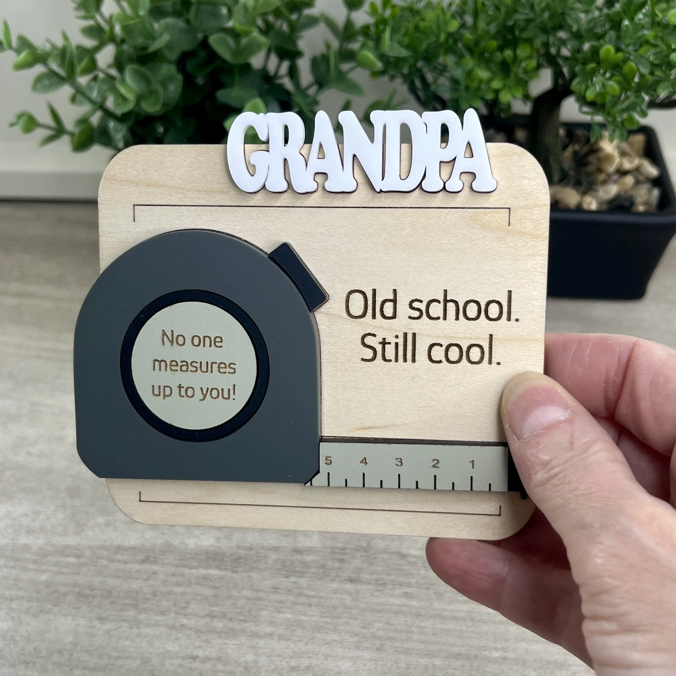 Handheld view of a maple wood grandpa magnet. Features charcoal grey 3D acrylic tape measure, raised white text header, and engraved quotes Old school Still cool and No one measures up to you.