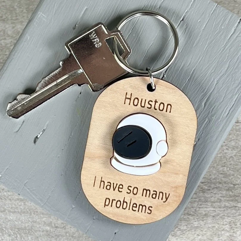 life style shot of laser cut maple keychain showing astronaut helmet graphic in raised matte white/black acrylic and engraved with "Houston I have so many problems".