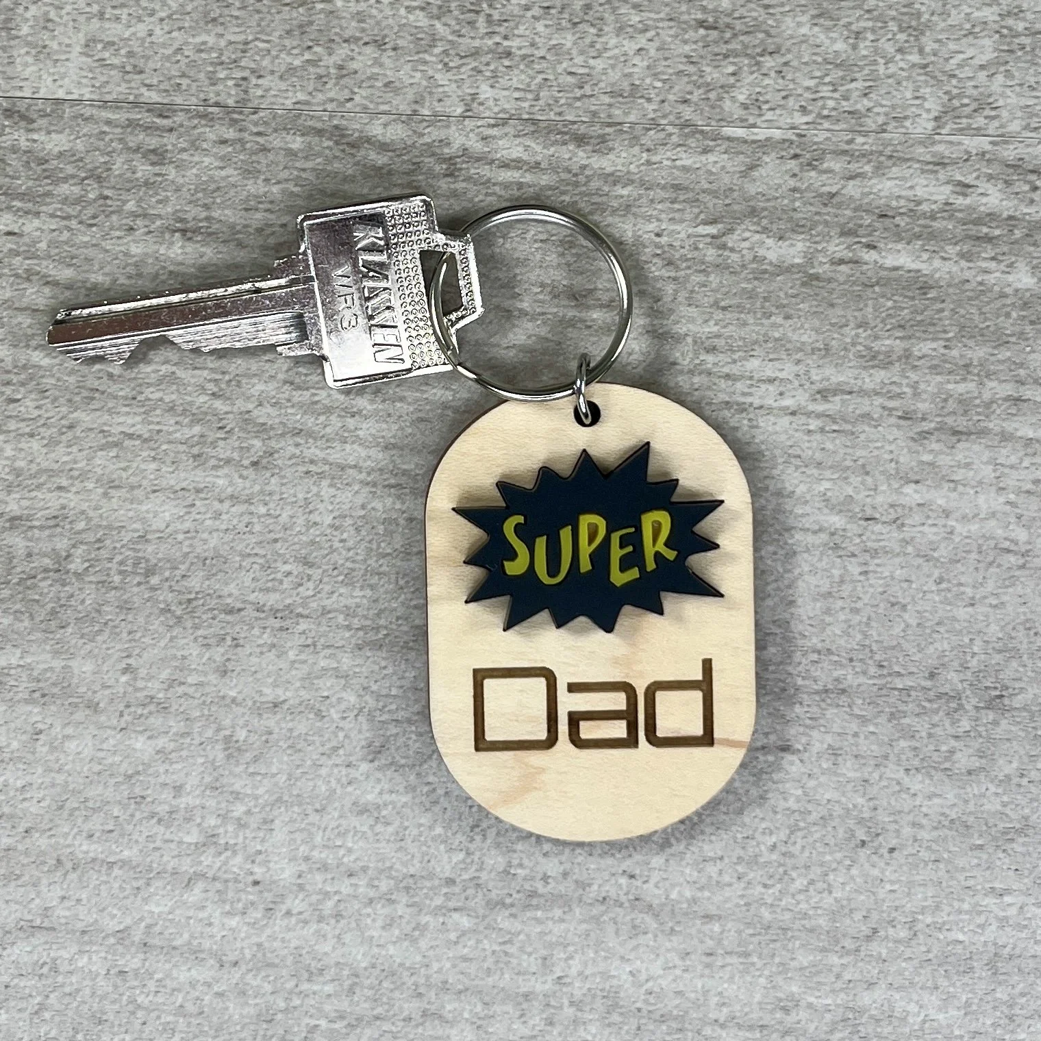 lifestyle shot of wood keychain black and yellow comic book burst shape reading Super with Dad engraved beneath.