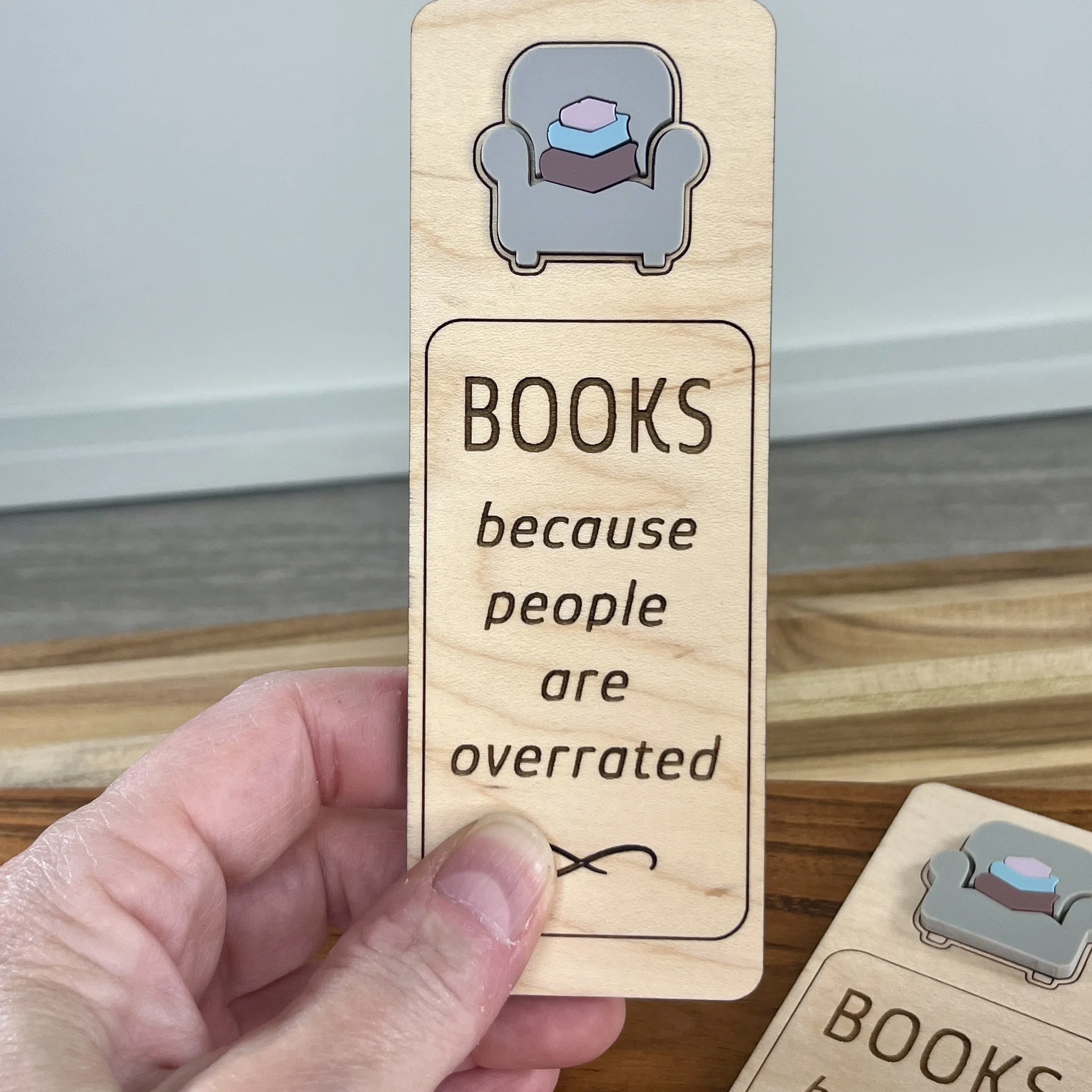 handheld maple bookmark featuring blue acrylic reading chair and stack of books embellishment engraved with the phrase "Books Because People Are Overrated".