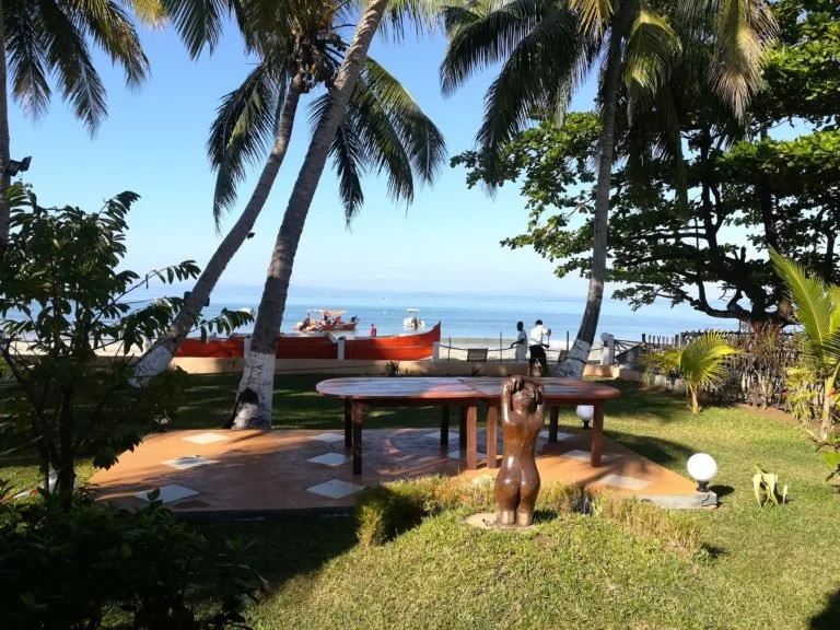 Spiaggia tropicale con palme, tavolo di legno, scultura di donna in primo piano, barche sulla riva e persone che camminano sulla spiaggia con mare e cielo azzurri sullo sfondo.