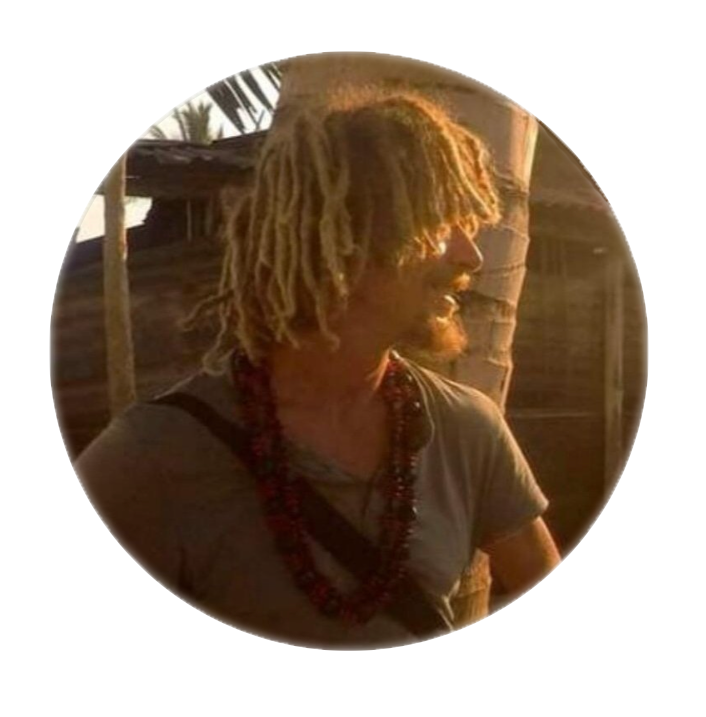 Uomo con dreadlocks e collane di fronte a uno sfondo di palme e una struttura di legno, illuminato dalla luce calda del tramonto.