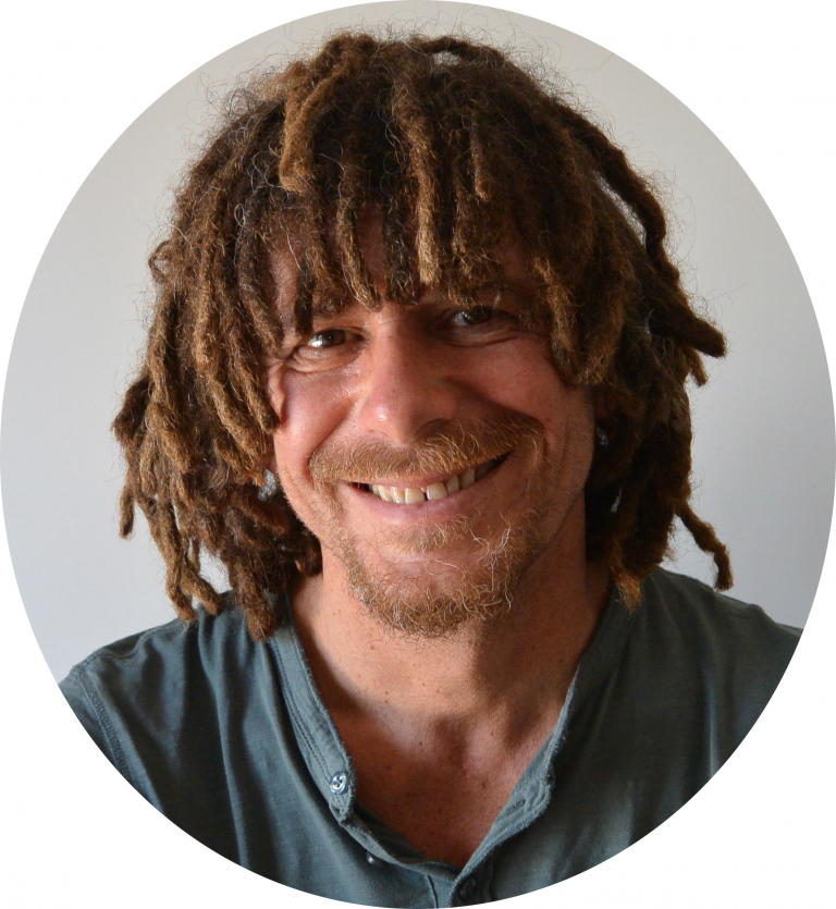Uomo sorridente con capelli in dreadlocks, barba, indossa una camicia grigia, sfondo bianco.