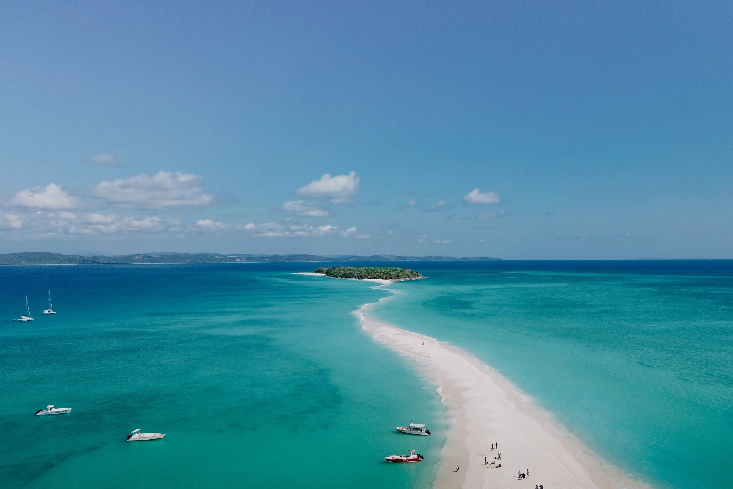 Spiaggia con sabbia bianca, l'acqua cristallina turchese, alcune barche e persone che camminano e si rilassano sulla riva, con un'isola verde sullo sfondo sotto un cielo soleggiato con alcune nuvole.