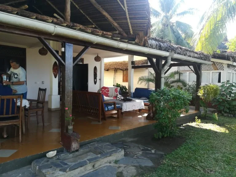 Veranda di una casa con mobili in legno, piante verdi e tetto di paglia, in un ambiente tropicale.