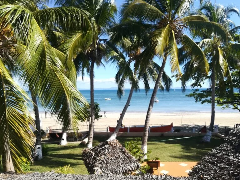 Spiaggia tropicale con palme, sabbia e mare blu, alcune barche in mare.