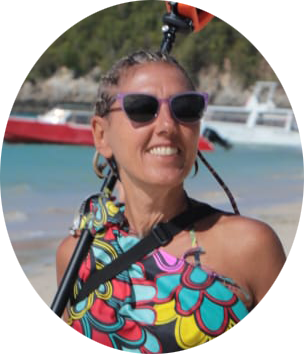 Una donna sorridente con occhiali da sole, in posa sulla spiaggia, con una canoa o sup sullo sfondo.