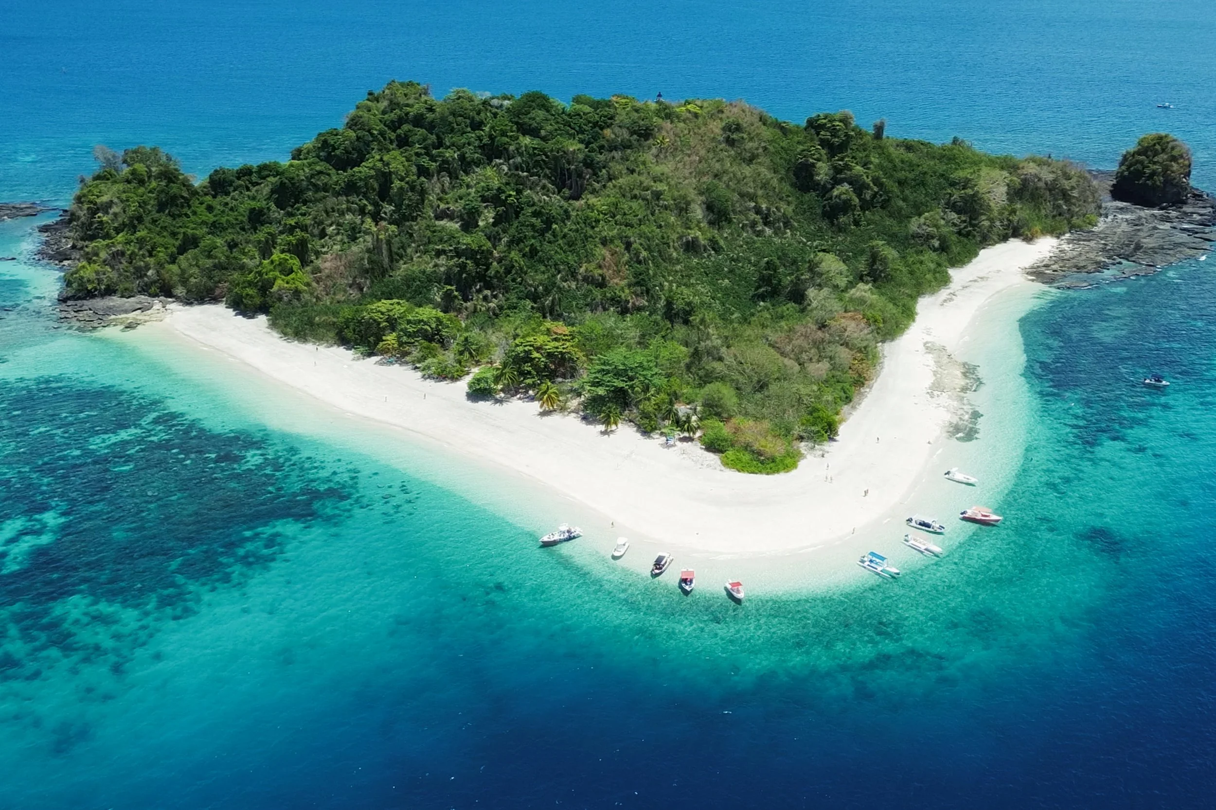 Isola tropicale con spiaggia di sabbia bianca, acqua turchese e vegetazione lussureggiante, con barche ormeggiate lungo la costa e acque cristalline