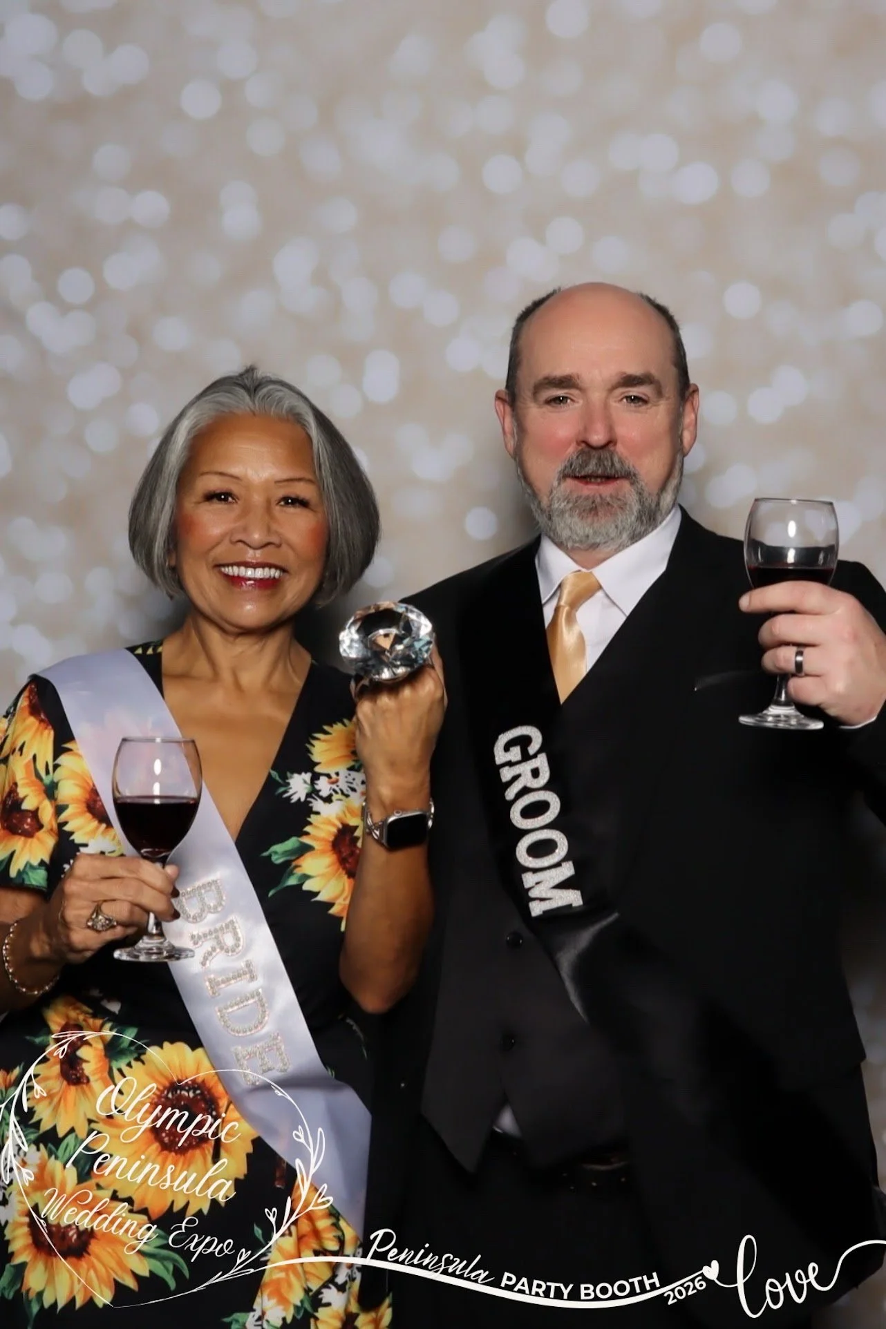 A smiling woman wearing a floral dress and a sash that reads "BRIDE" and a man in a tuxedo with a sash that reads "GROOM" holding glasses of red wine at a wedding expo.