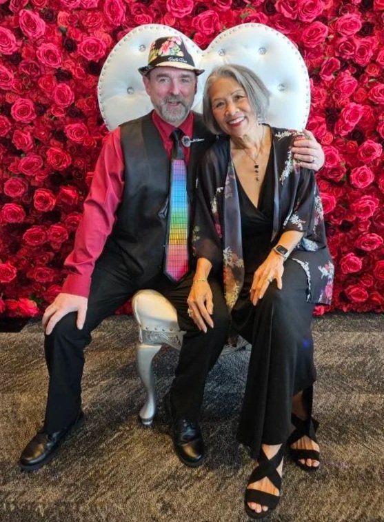A man and a woman sitting together in front of a background of red roses and a white heart-shaped decoration. The man is wearing a colorful hat, a red shirt, a black vest, and a rainbow tie, with his arm around the woman's shoulder. The woman is wearing a black dress with a floral shawl, black sandals, and has gray hair. They are smiling and posing for the photo.