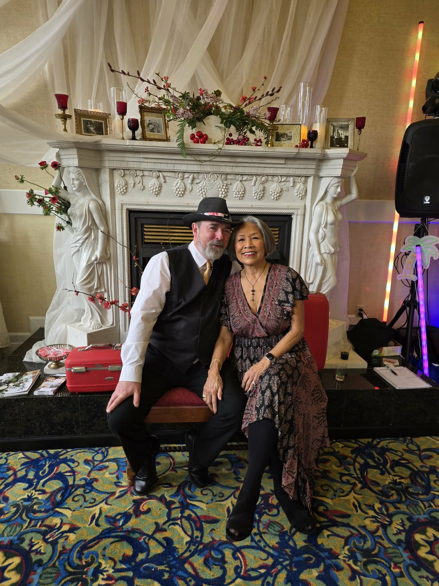 Two people sitting on a bench in front of a decorated fireplace. The man is wearing a black hat, white shirt, and black vest, while the woman is dressed in a patterned gown. They are smiling and posing for the camera.