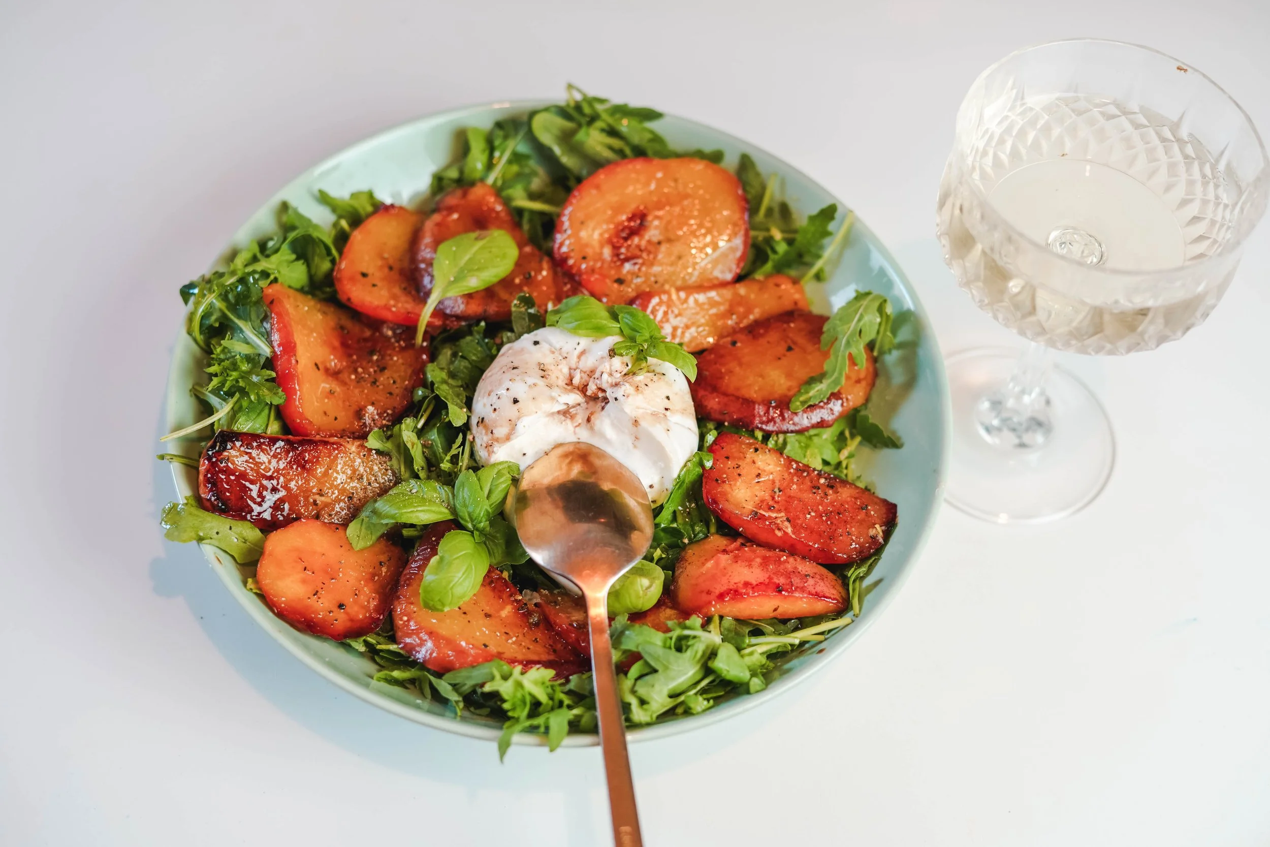 A salad with roasted peaches, fresh greens, basil, a poached egg, and black pepper in a blue bowl, with a glass of water nearby.