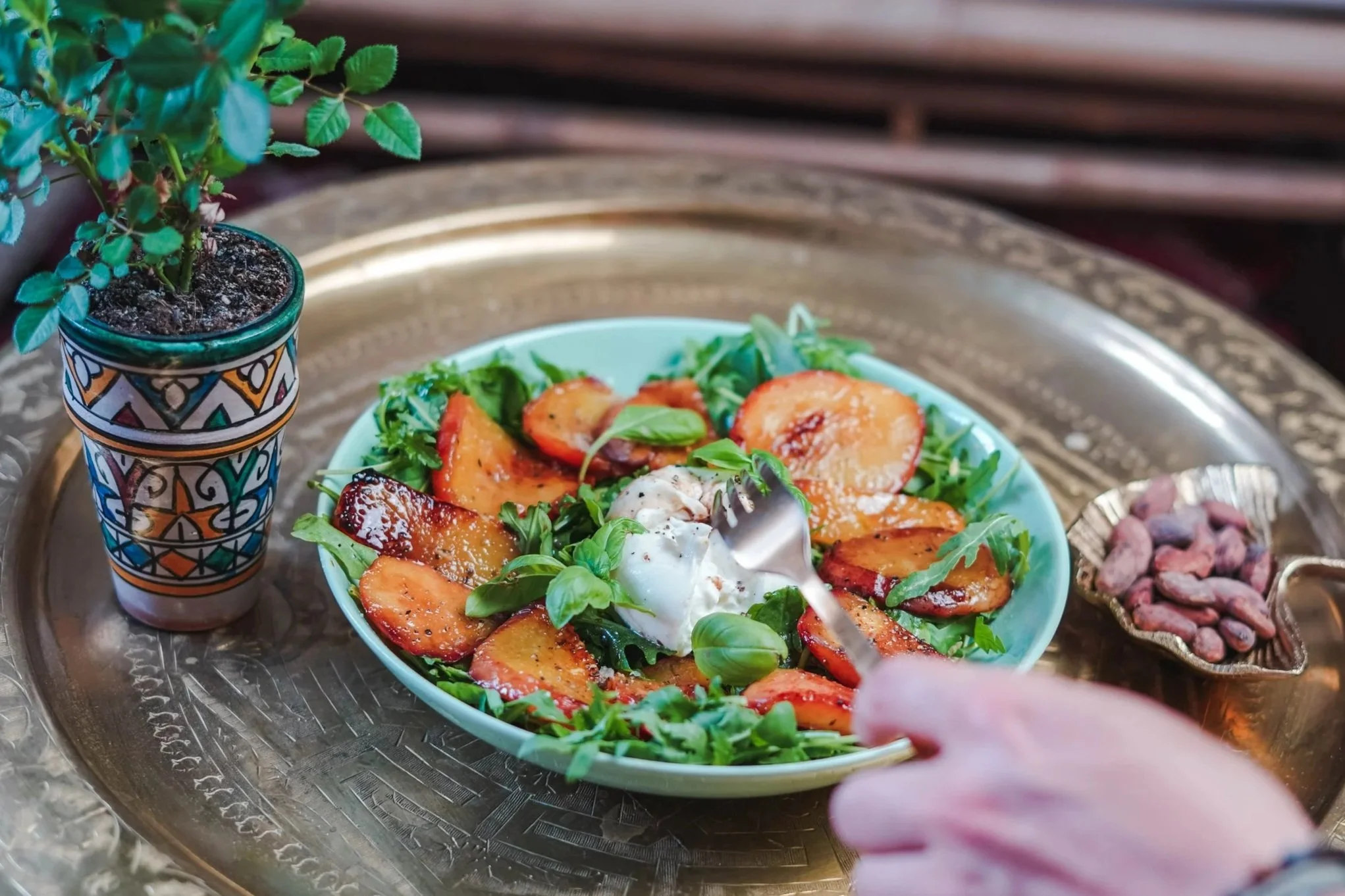 a bowl of salad with grilled peaches, greens, and burrata cheese on a gold tray, with a hand holding a fork, next to a potted plant and small dish of nuts.