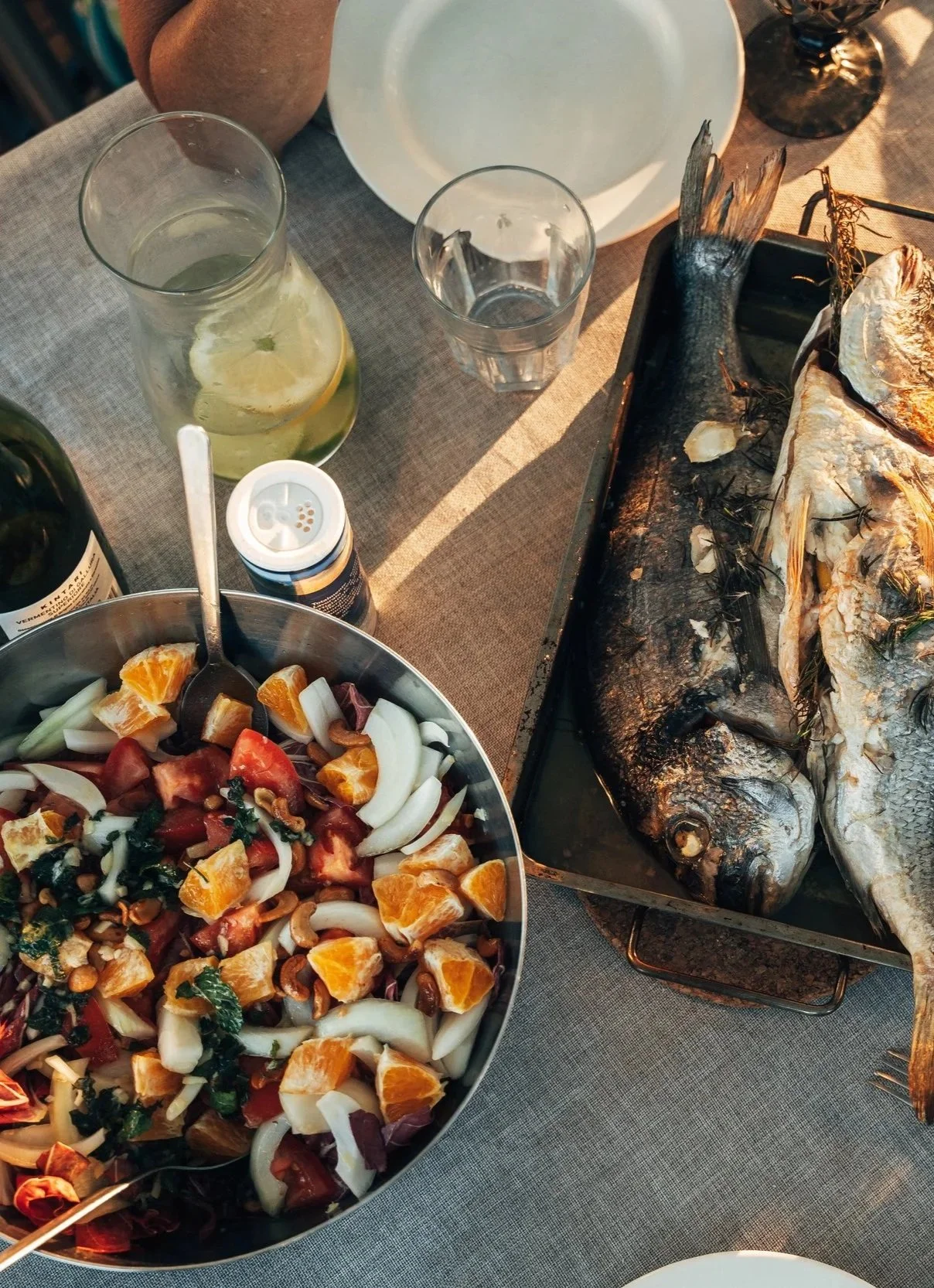 Grilled fish on a baking sheet, a large colorful salad with oranges, tomatoes, onions, and pine nuts in a bowl, and drinks with lemon slices on a dining table.