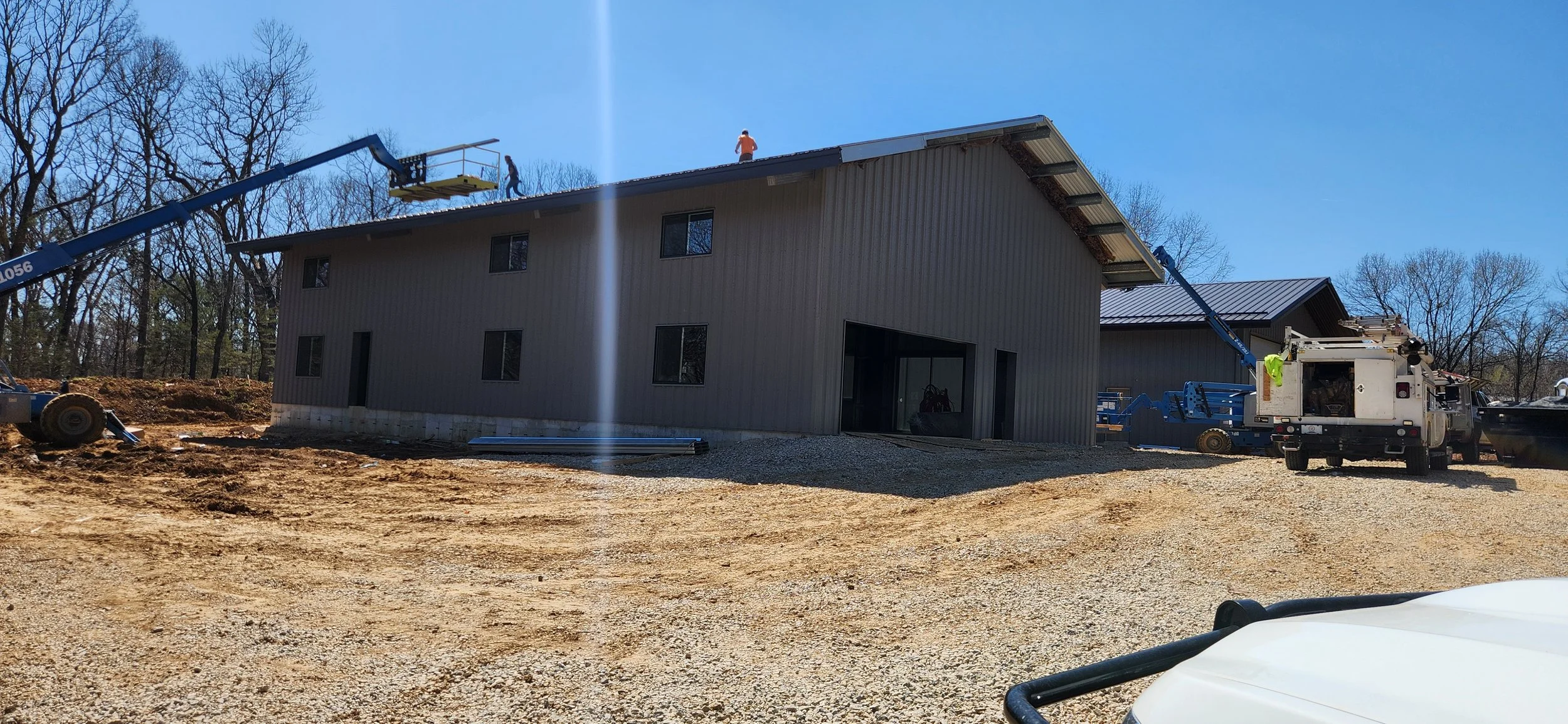Construction workers on a building roof with a telescopic boom lift, building exterior, gravel ground, blue sky, leafless trees in the background.