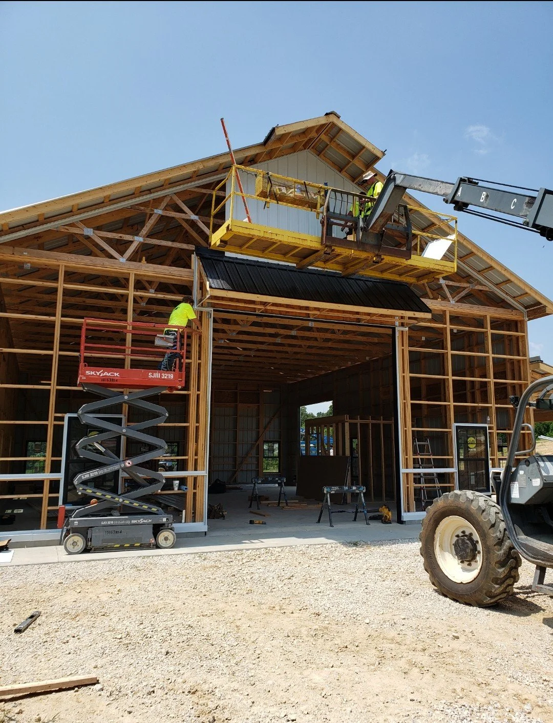 Construction workers building a wooden house, with one working on the ground and another on an elevated platform, under a clear blue sky.