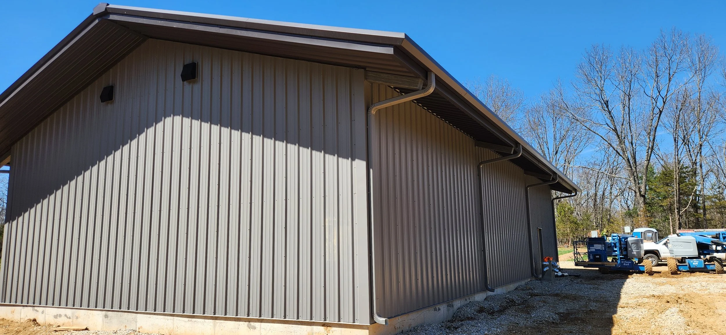 Side view of a large metal building with vertical siding, a sloped roof, and outdoor lighting fixtures, with construction equipment and trees in the background on a clear day.