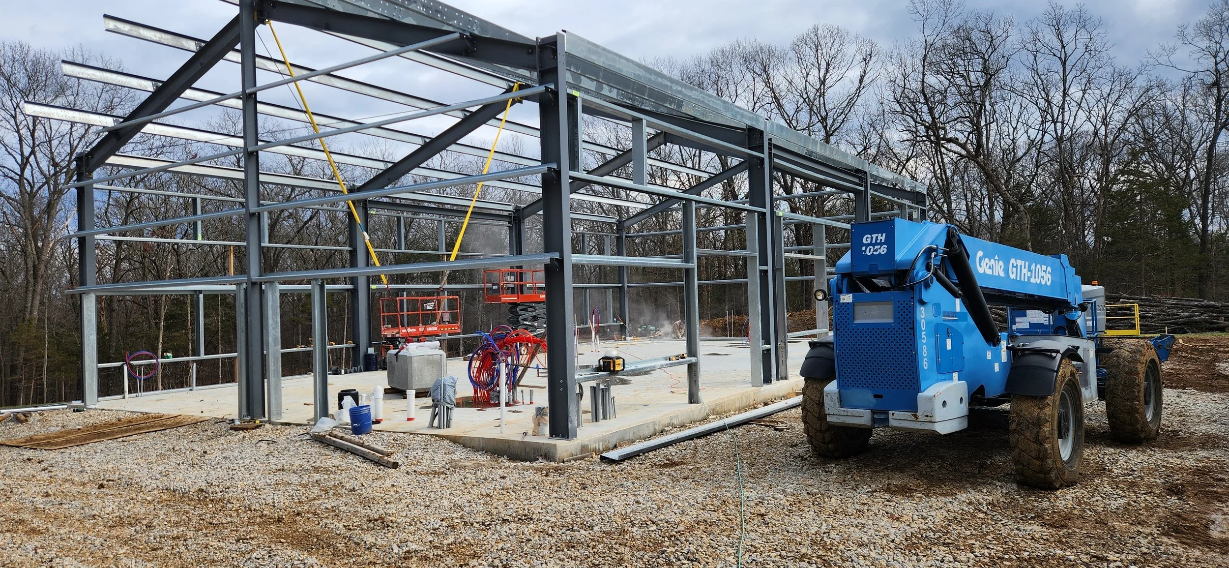 Construction site with a partially built metal structure, concrete foundation, construction equipment, and machinery, including a blue Genie GTH-1056 telehandler, surrounded by gravel and trees in the background.