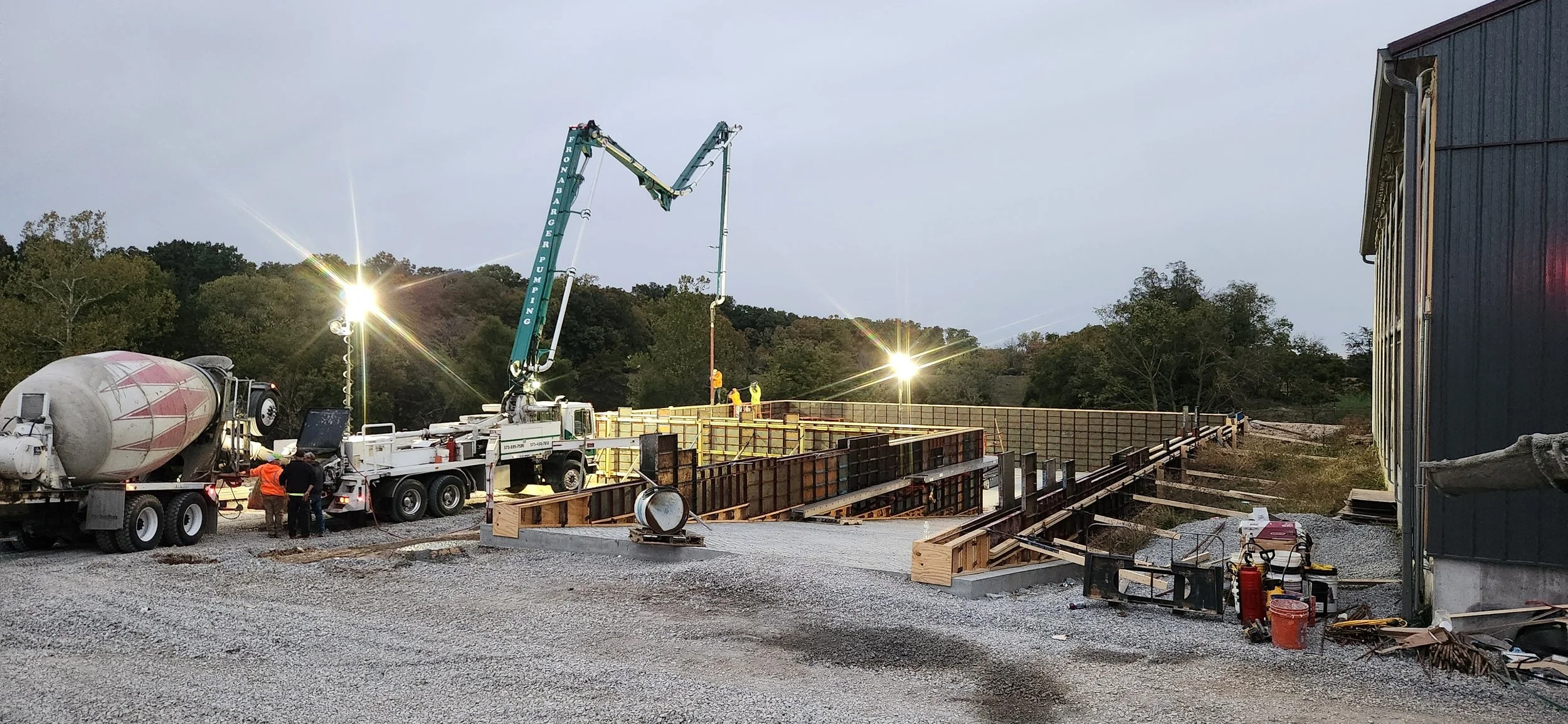 Construction site with a concrete mixer truck, a crane pouring concrete into formwork, working workers, and construction materials, under a cloudy sky.