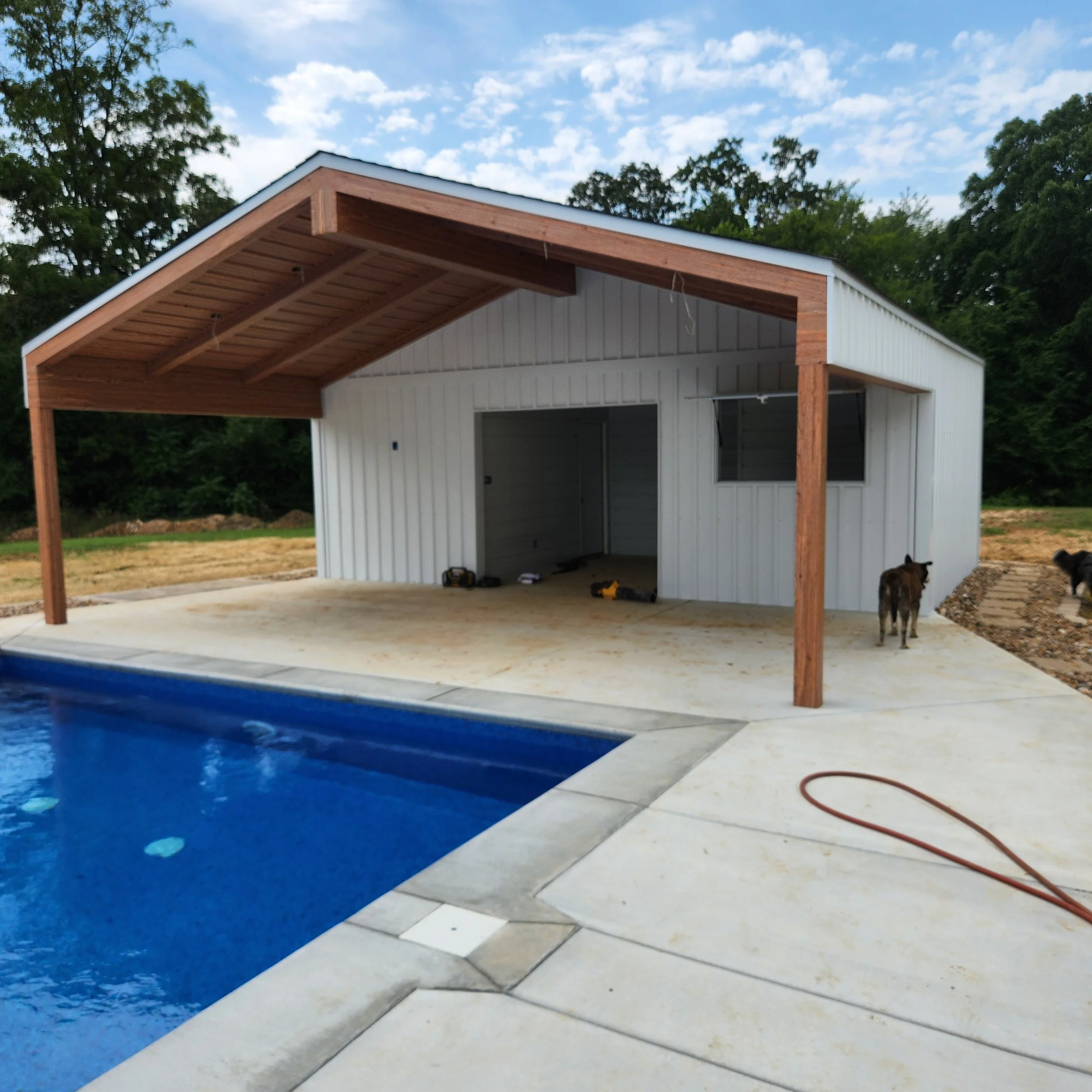 A backyard with a swimming pool and a newly built pool house with a covered patio area, construction tools on the ground, and dogs nearby.