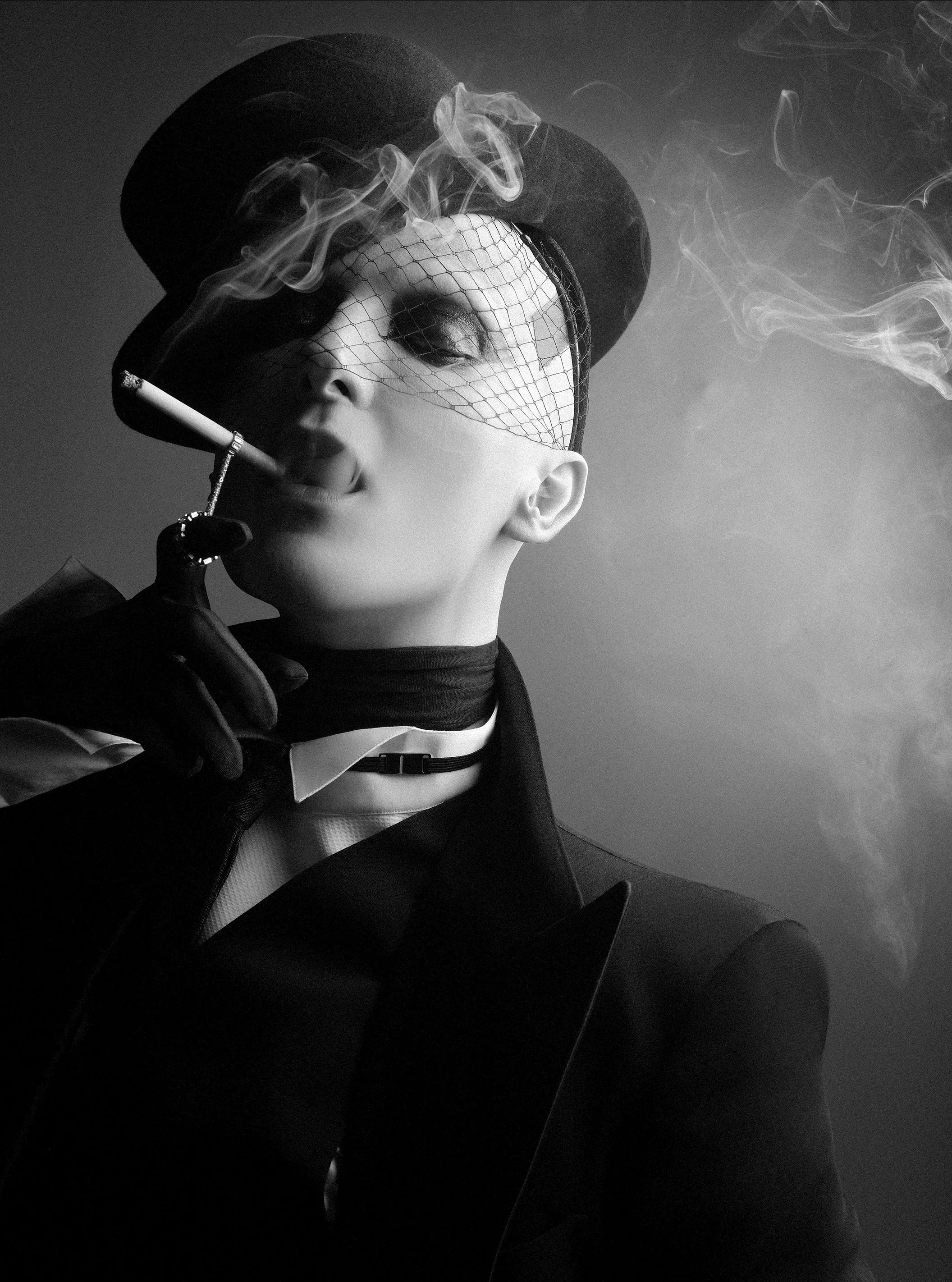 Black and white portrait of a person with short hair, wearing a hat with netting, a choker, and a tailored jacket. They are smoking a cigarette, with smoke swirling around their face.